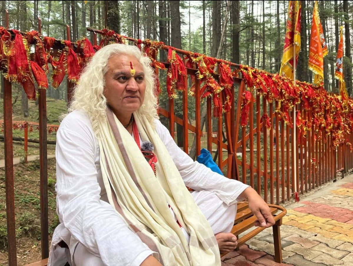 Manoj Dhyani, white-haired and in white, seated in front of a railing hung with red chunris in a deodar forest